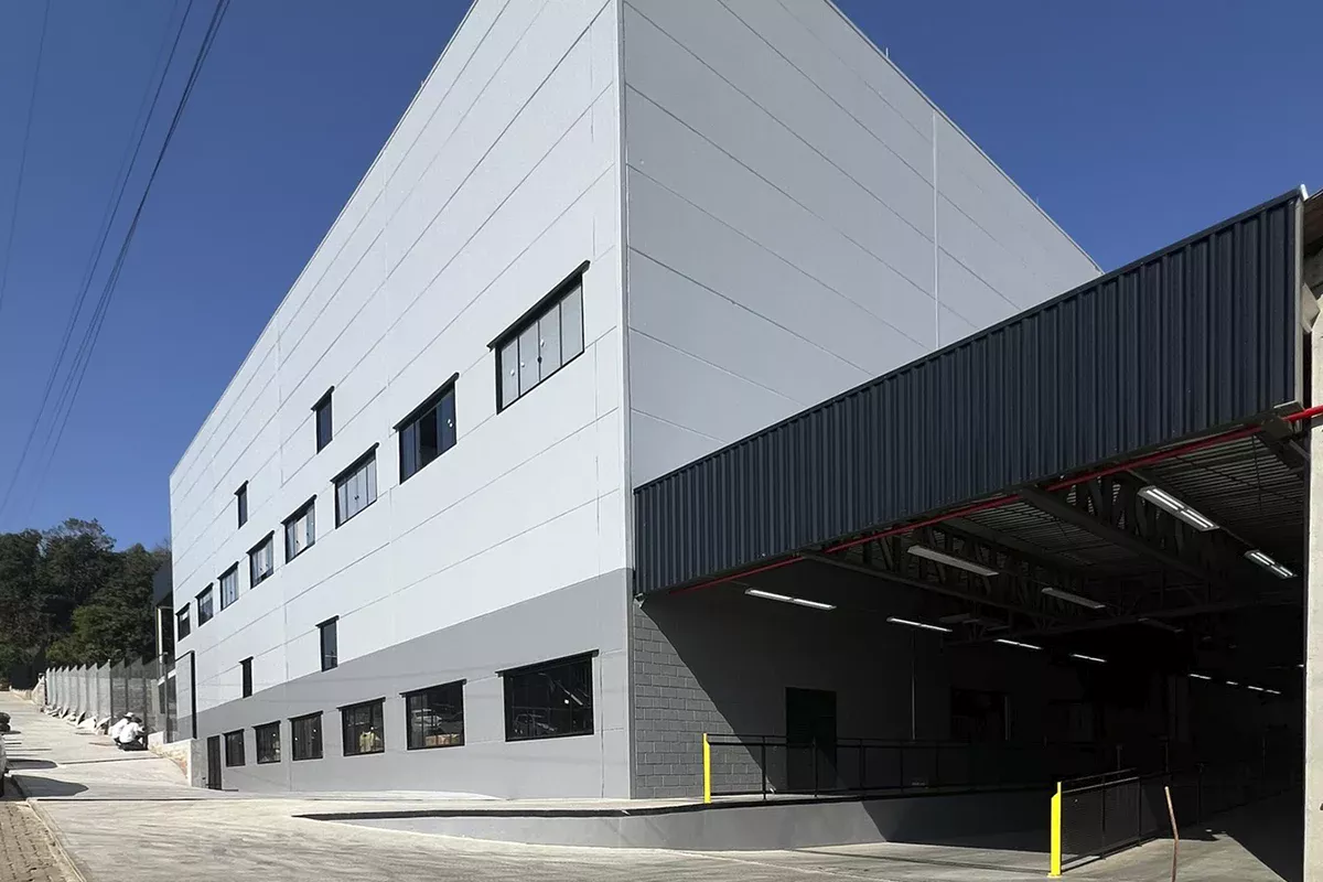 Obras casa Dass - Fachada lateral de galpão industrial moderno, estrutura metálica aparente, paredes cinza claro e amplas janelas, evidenciando Soluções em Comunicação e excelência operacional.