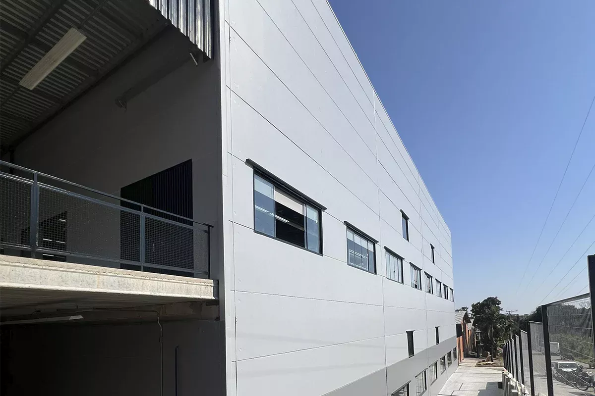 Obras casa Dass - Detalhe da fachada lateral do galpão industrial, múltiplas janelas alinhadas, plataformas cobertas e design funcional, evidenciando foco em resultados mensuráveis.