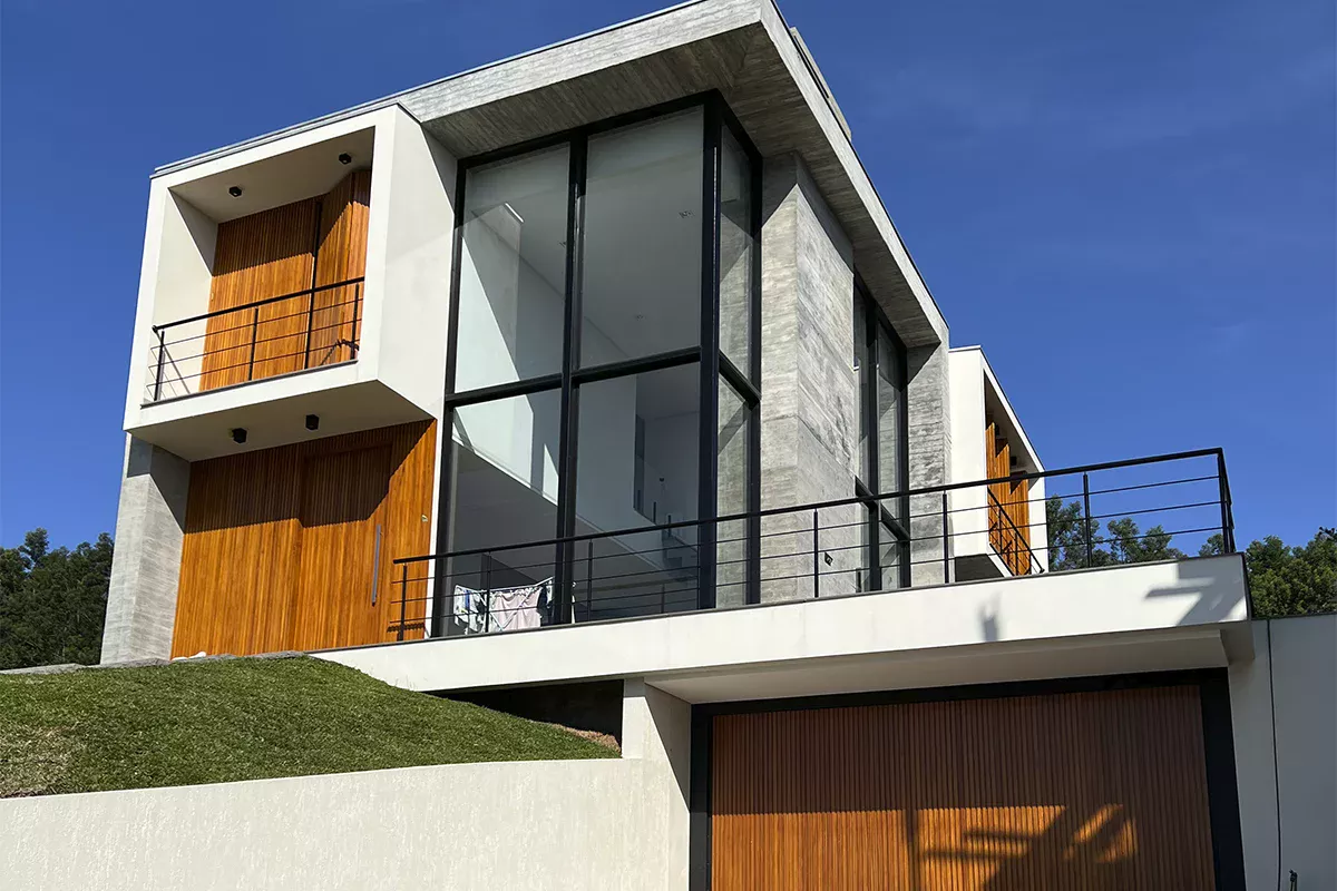 Foto de uma casa contemporânea com fachada em concreto aparente, grandes janelas de vidro e detalhes em madeira, vista de um ângulo frontal à esquerda, gramado verde à frente.