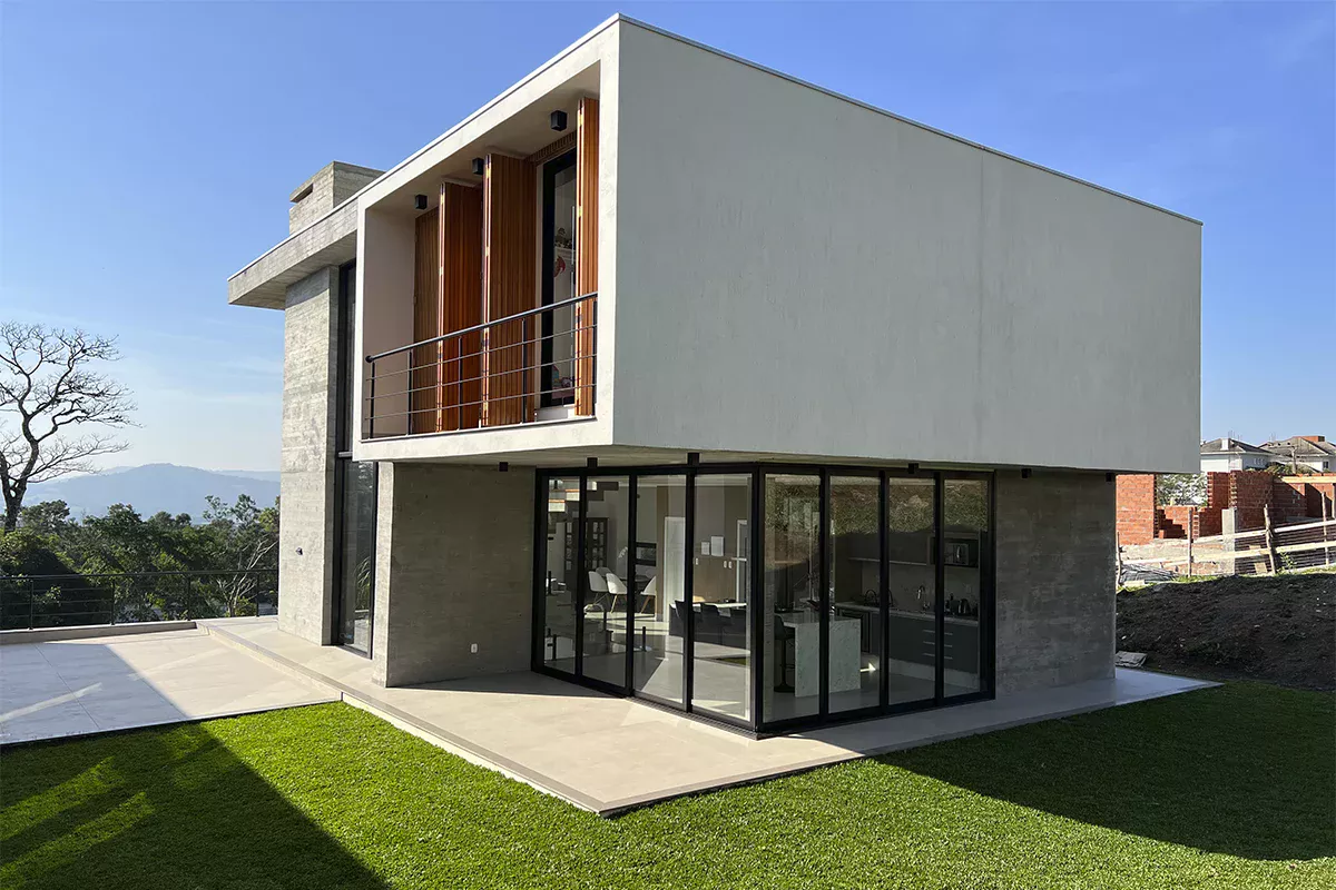 Foto de residência moderna, com fachada de concreto aparente, janelas de vidro do chão ao teto e detalhes em madeira, fotografada em perspectiva frontal e lateral, realçando o jardim e a iluminação natural.