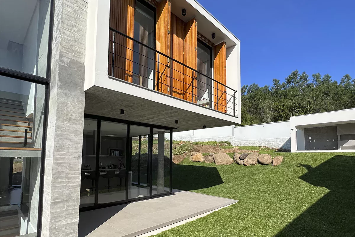 Foto de casa de arquitetura moderna, com fachada de concreto, grandes áreas envidraçadas e elementos em madeira, vista em ângulo lateral direito, com céu azul ao fundo e gramado ao redor.