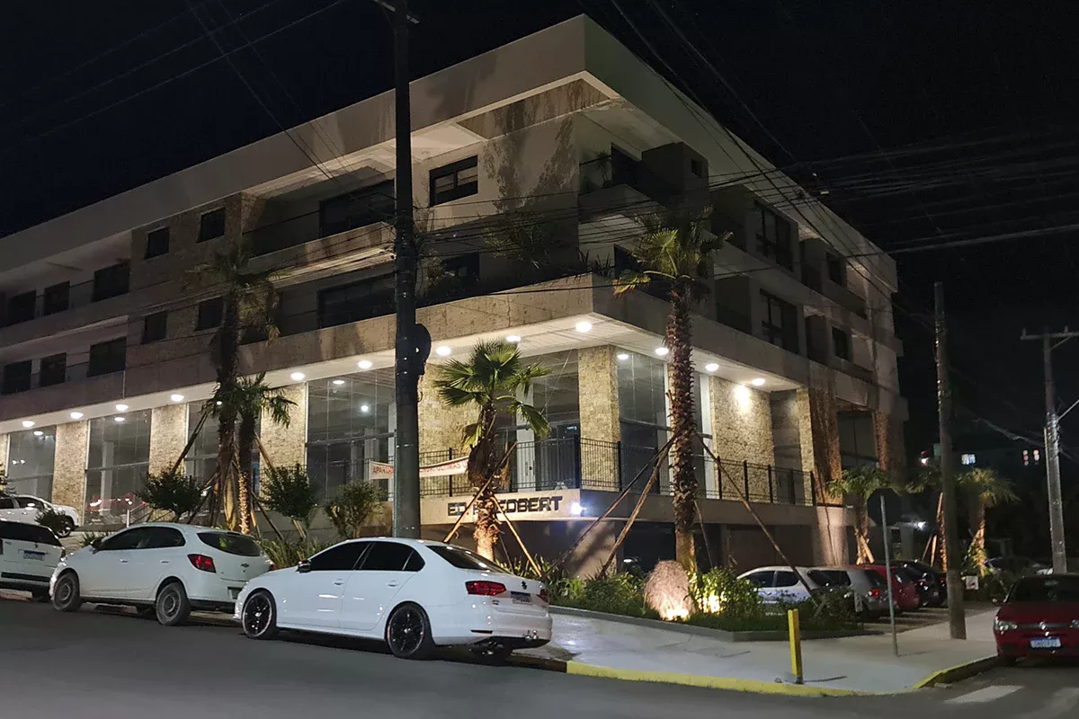 Obras casa Friedbert - Vista noturna de edifício comercial, iluminação estratégica destacando arquitetura moderna, paisagismo externo e estacionamento com veículo, enfatizando inovação e experiência diferenciada.