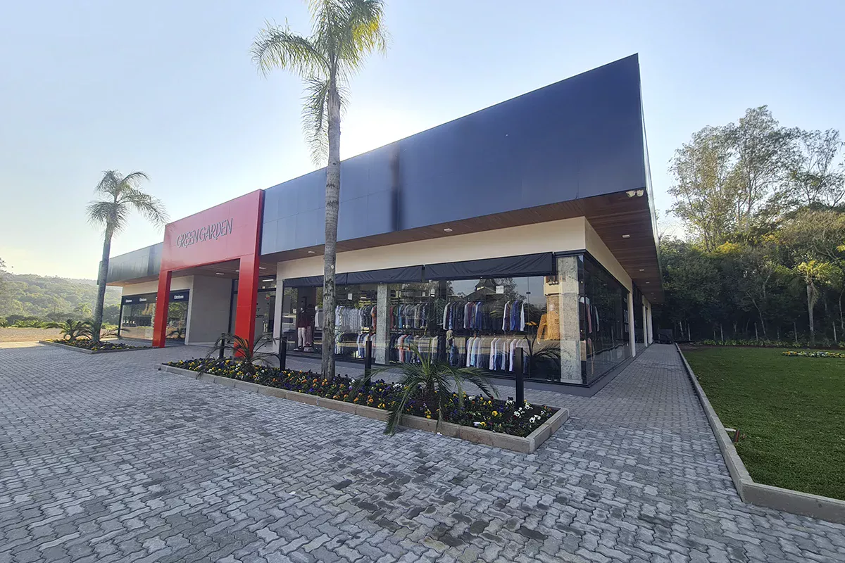 Obras casa Green Garden - Fachada de centro comercial contemporâneo, com linhas retas e acabamento em painel metálico azul e vermelho, vitrines amplas e calçamento em bloco intertravado, evidenciando soluções em comunicação para o varejo.