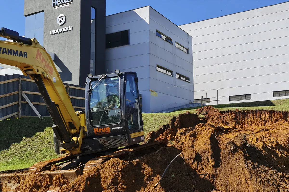 Obras casa Hexus - Obra em andamento, escavadeira atuando em terraplanagem ao lado de prédio comercial moderno, refletindo expertise técnica e gestão comercial eficiente.