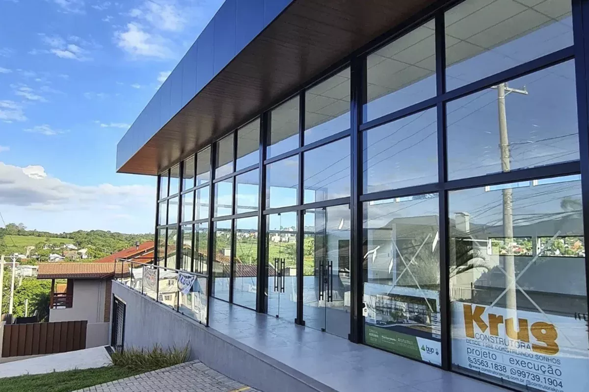 Obras casa Klauck - Perspectiva angular do galpão, grandes janelas envidraçadas sob marquise metálica, área externa pavimentada e projeto paisagístico, simbolizando ambientes funcionais, soluções resolutivas e eficiência operacional.