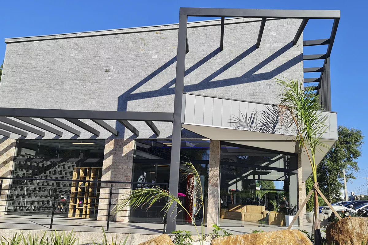 Obras casa Me - Entrada de centro comercial contemporâneo com pergolado metálico, paisagismo integrado e elementos arquitetônicos inovadores, transmitindo atendimento resolutivo e foco em experiência do cliente.