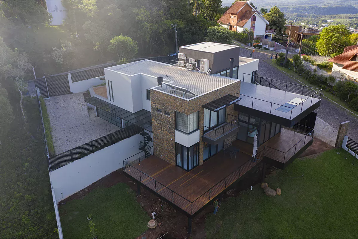 Obras casa MR - Vista aérea de residência contemporânea de dois pavimentos, com terraço em madeira, ampla área verde e muro de proteção, evidenciando inovação arquitetônica e soluções personalizadas.