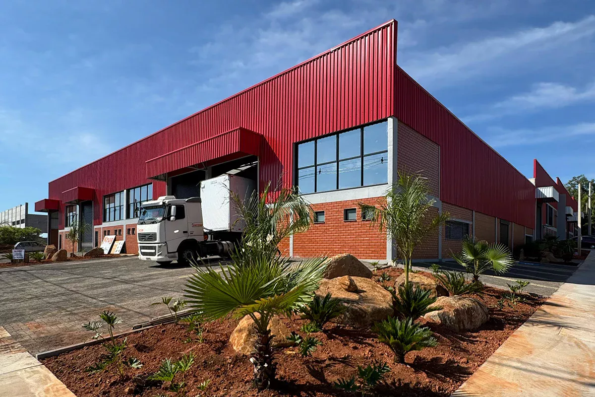 Obras casa Pavilhão Comercial - Vista frontal do galpão industrial vermelho com paisagismo, rampas de acesso e áreas externas organizadas, transmitindo excelência técnica e alinhamento estratégico.