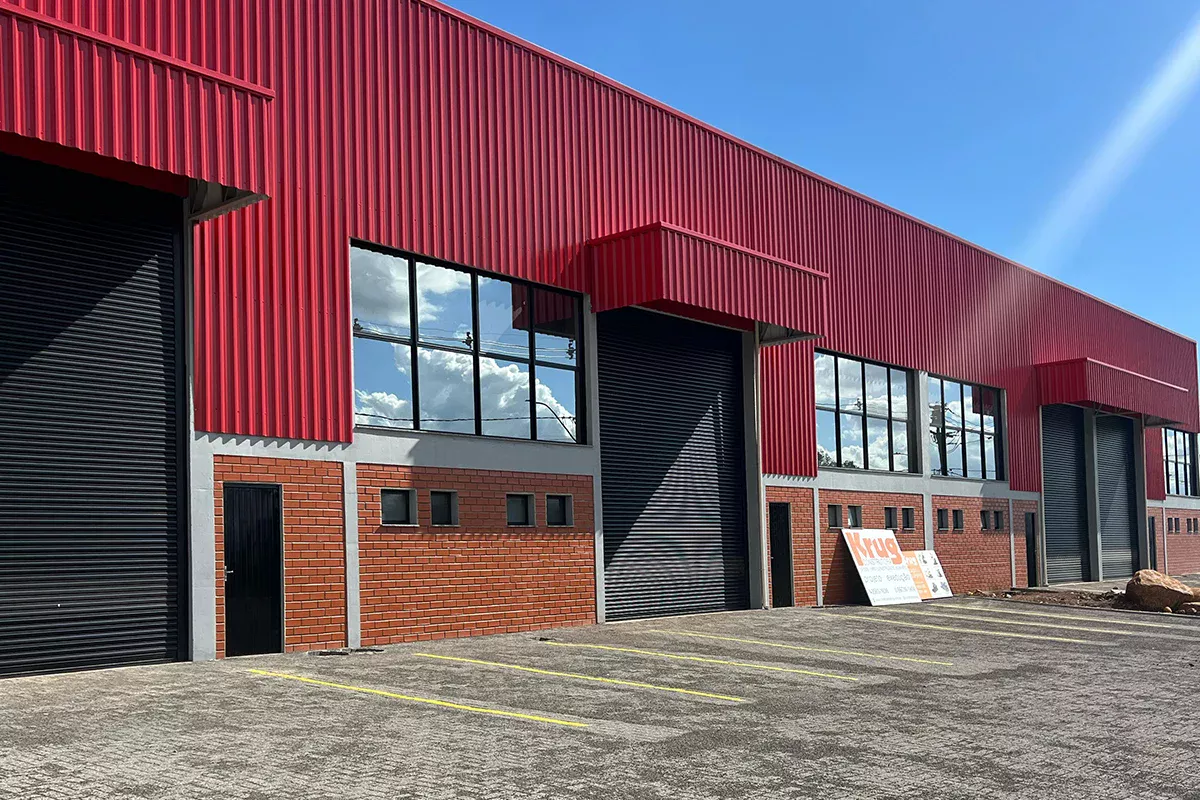 Obras casa Pavilhão Comercial - Galpão industrial com fachada em chapa metálica vermelha e alvenaria à base, entradas amplas e piso pavimentado, evidenciando inovação construtiva e Soluções em Comunicação voltadas ao segmento corporativo.